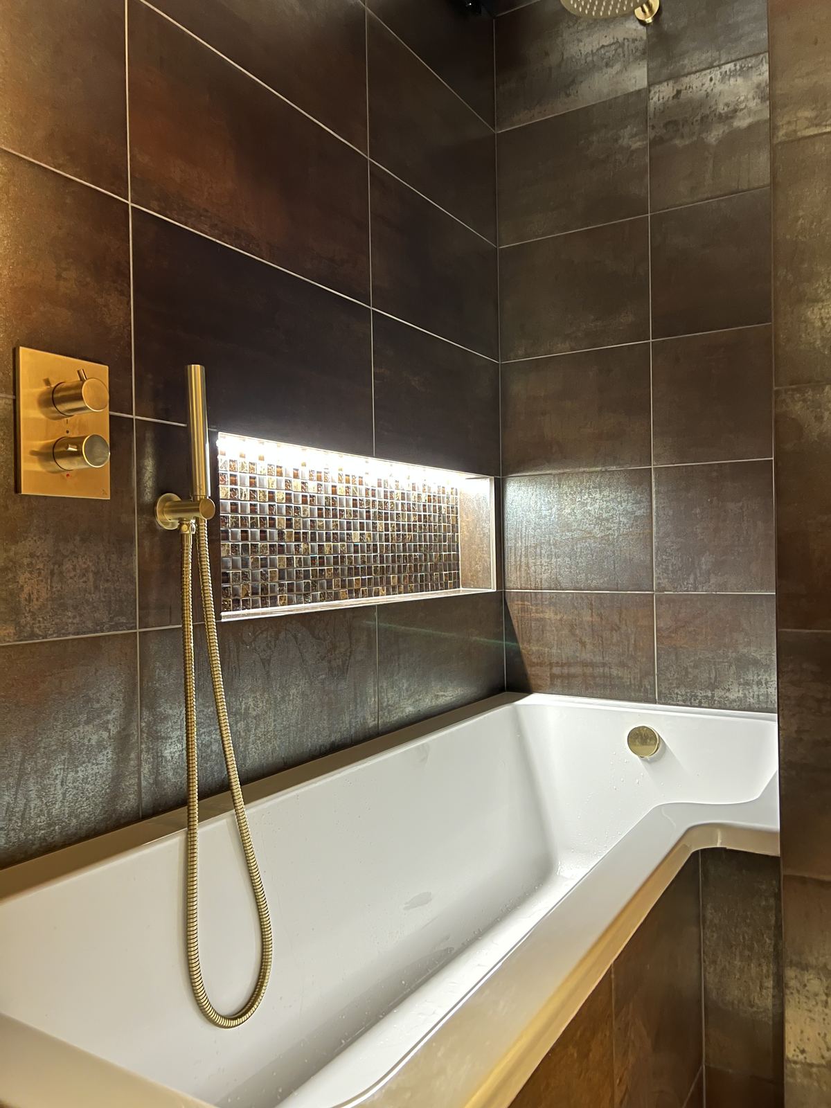Brushed brass shower beside a back-lit mosaic niche